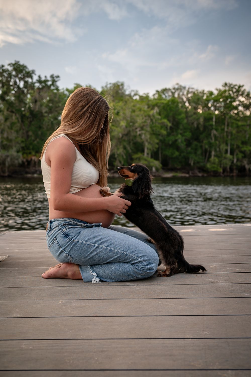 Pregnant woman in white crop top and jeans sitting on wooden dock with black dachshund by serene lake.