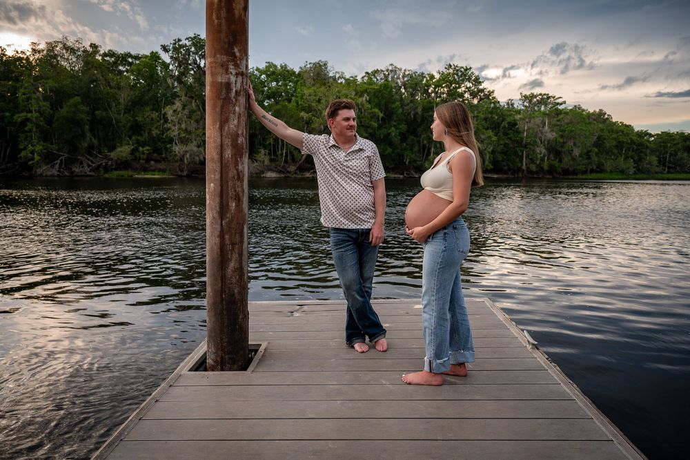 Couple sharing a tender moment on a wooden dock by a serene lake at sunset, woman visibly pregnant.