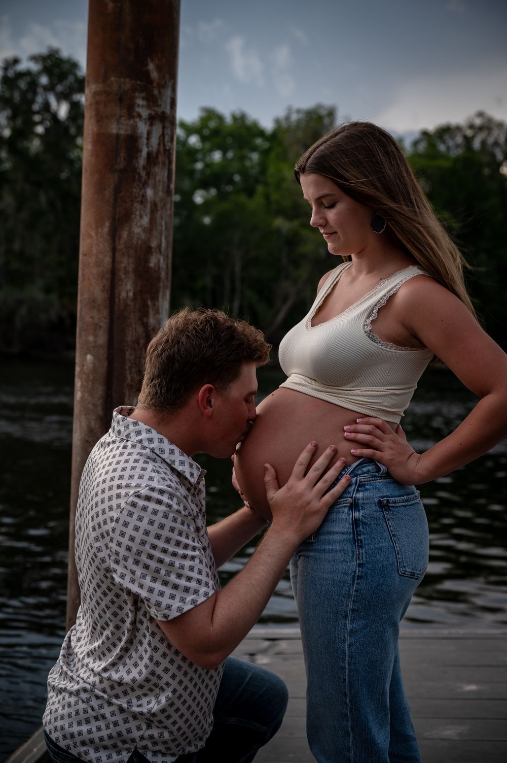 Expectant father kneeling to kiss pregnant woman's bare belly on a lakeside dock at dusk.