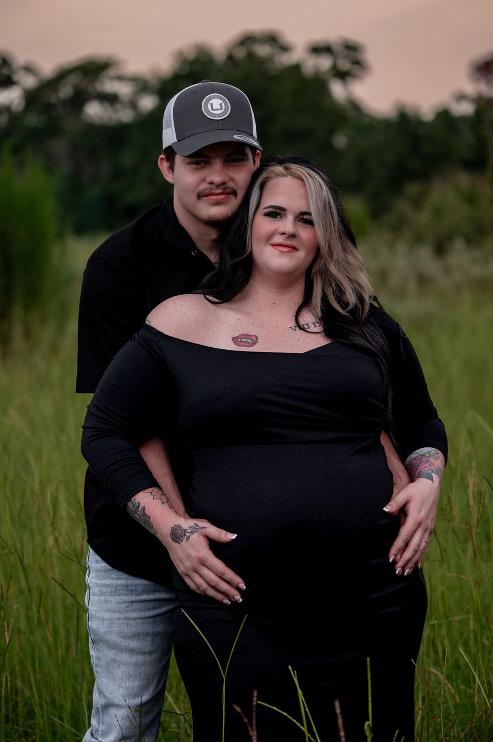 Couple posing in a green field at dusk, woman in black off-shoulder dress showing baby bump, man in gray trucker hat behind her.