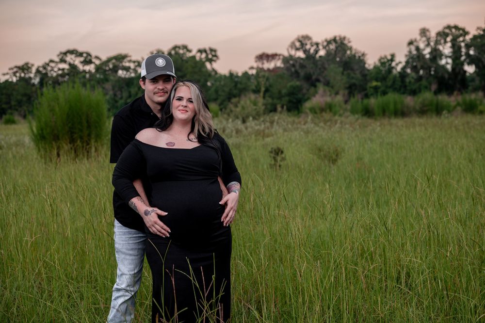 Couple posing in green field at dusk, woman in black maternity dress cradling baby bump, man in black shirt and cap.