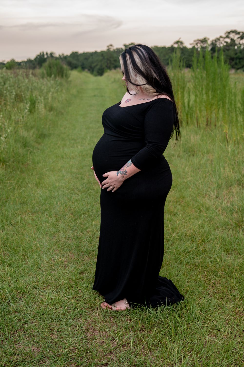 Pregnant woman in black maxi dress standing barefoot in green field, cradling baby bump during outdoor maternity photo session.