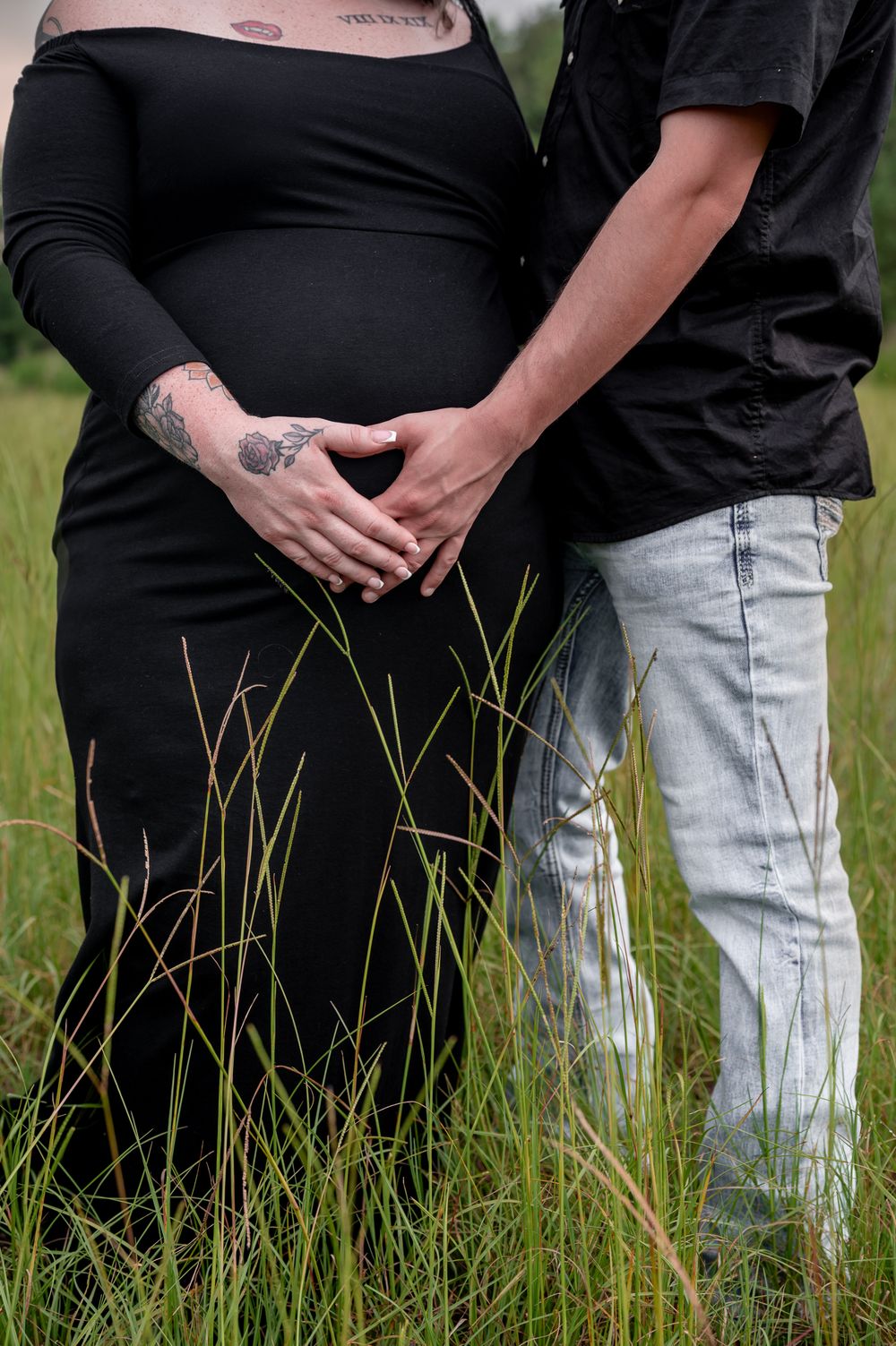 Pregnant couple in black outfits standing in tall grass, forming a heart shape with their hands over the baby bump.