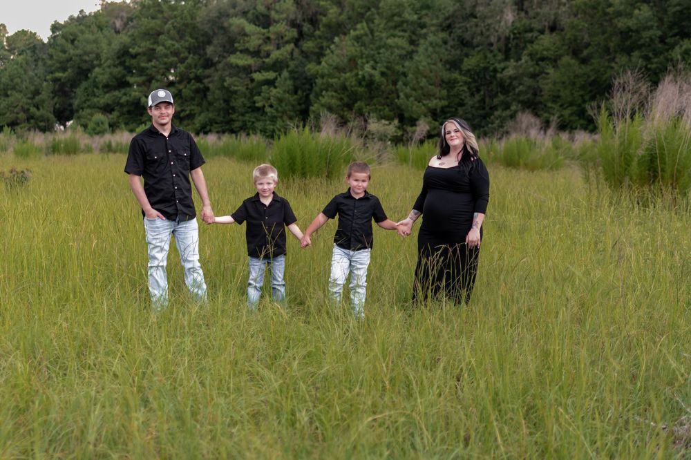 Family of four in black outfits holding hands in a lush green meadow, with a pregnant mother on the right.