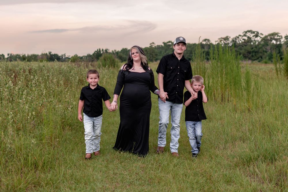 Pregnant woman in black gown poses with family in green field at sunset, all wearing matching black outfits.
