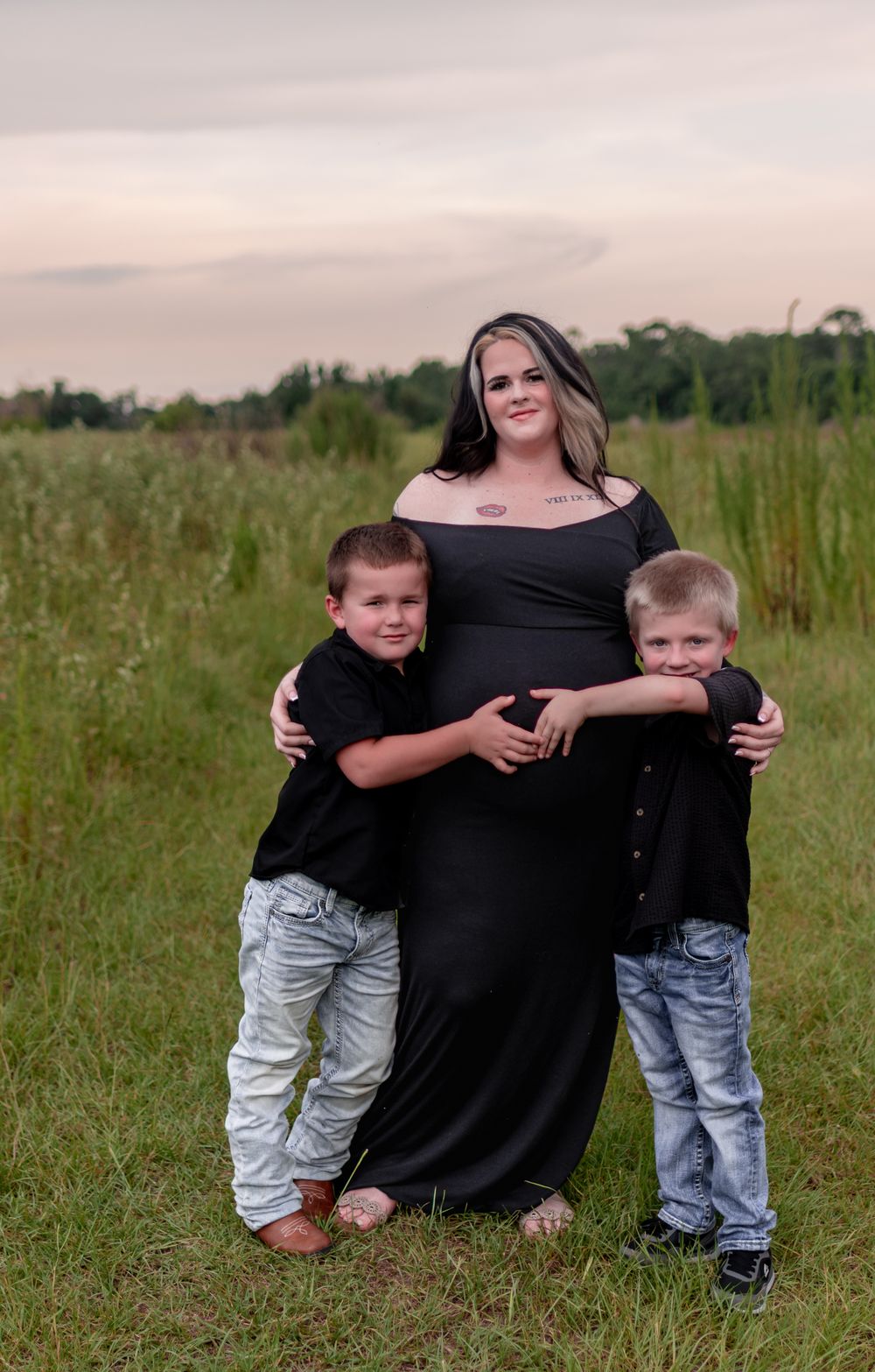 Pregnant woman in black maternity gown poses outdoors in a field with two young boys in matching black shirts and jeans.