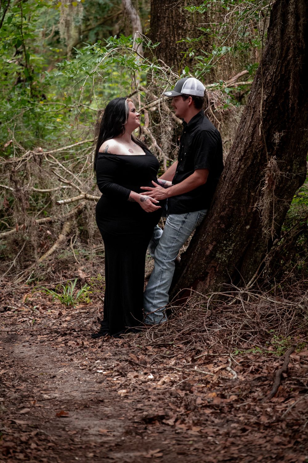 Pregnant couple holding baby bump together in a forest, woman in black maternity gown, man in black shirt and cap.