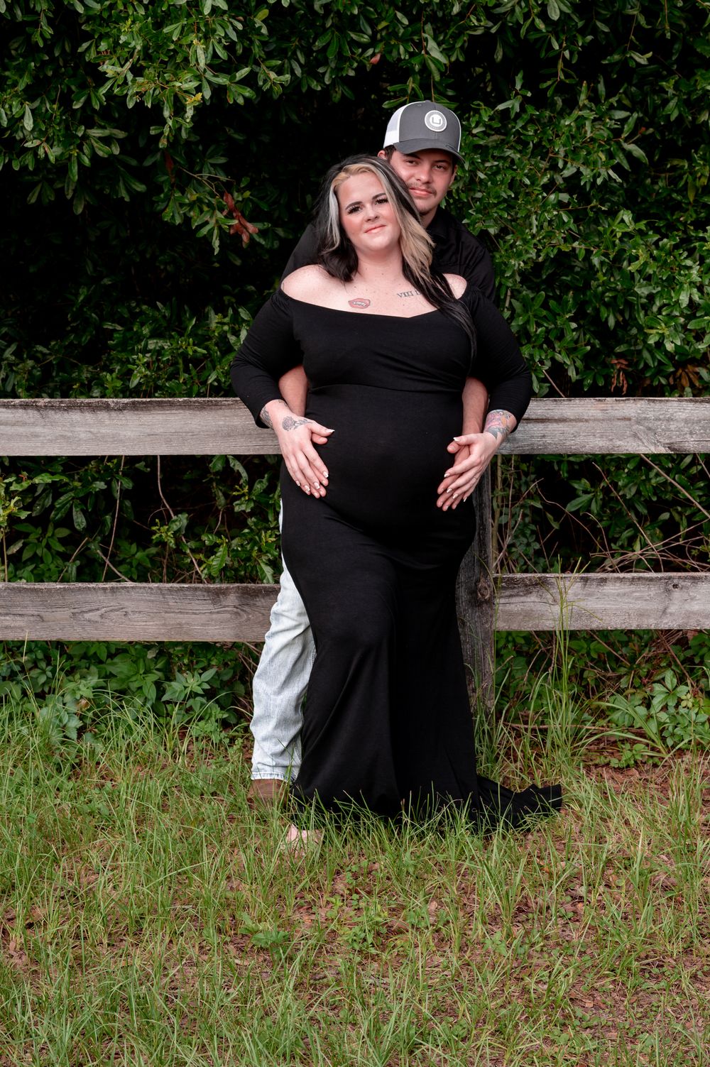 Pregnant woman in black off-shoulder gown poses with partner by rustic fence surrounded by lush greenery.