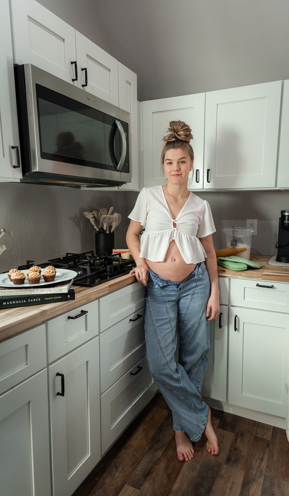 Pregnant woman in white crop top and jeans leaning against white kitchen counter with muffins nearby.