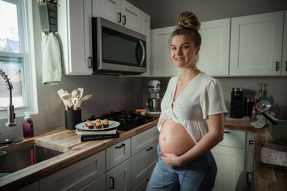 Smiling pregnant woman in white top and jeans cradling baby bump in modern kitchen with cupcakes on counter.