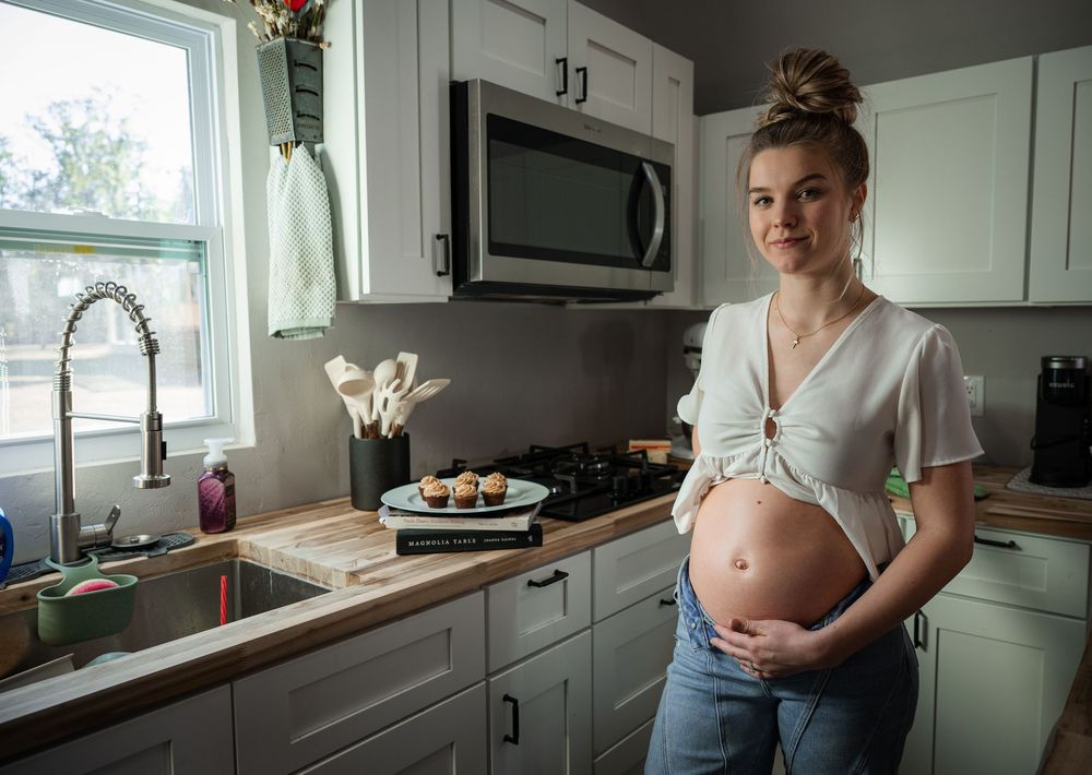 Pregnant woman in white crop top and jeans cradling baby bump in modern white kitchen with cupcakes on counter.