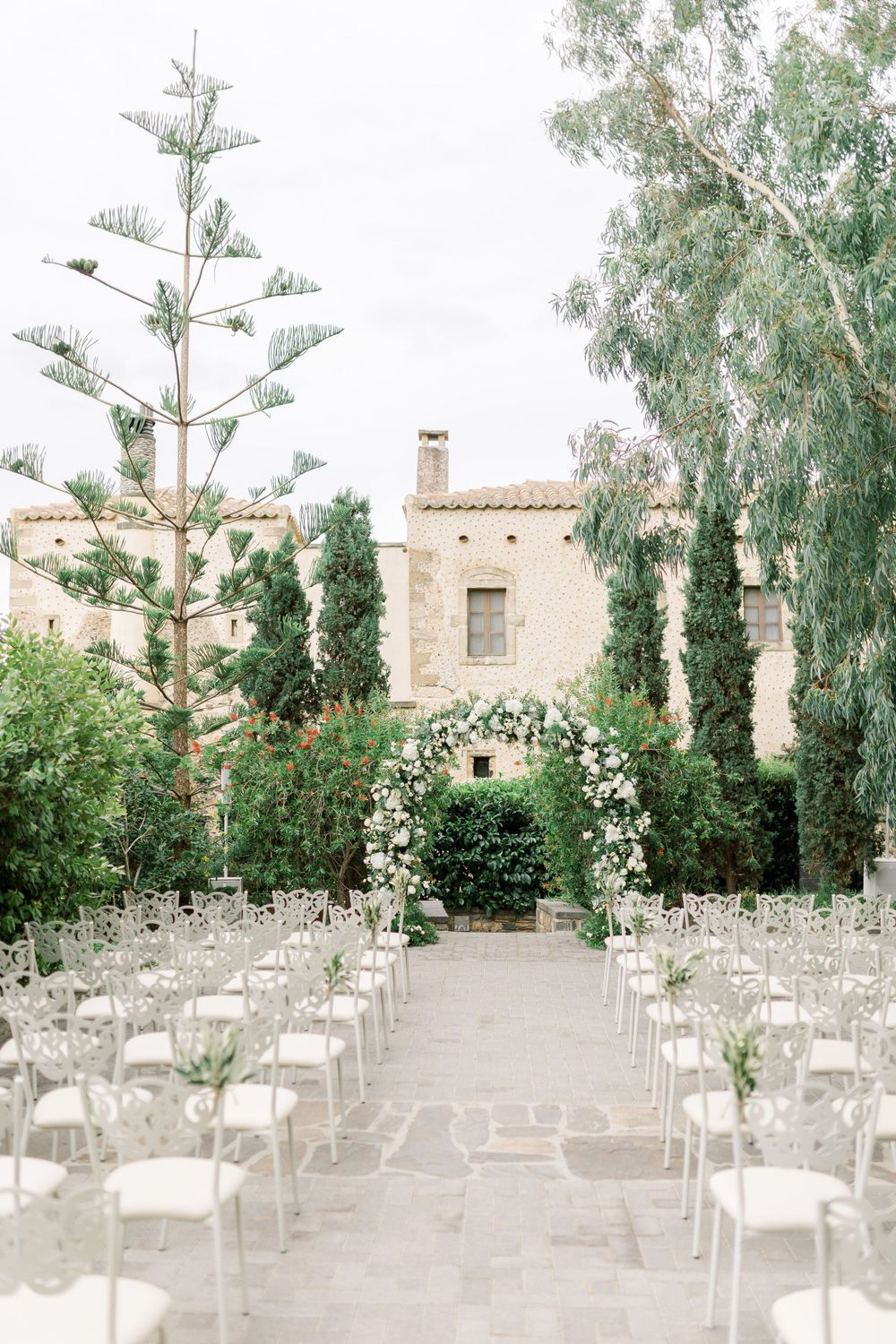 Outdoor wedding ceremony setup with white chairs and floral arch framing a Mediterranean style building.