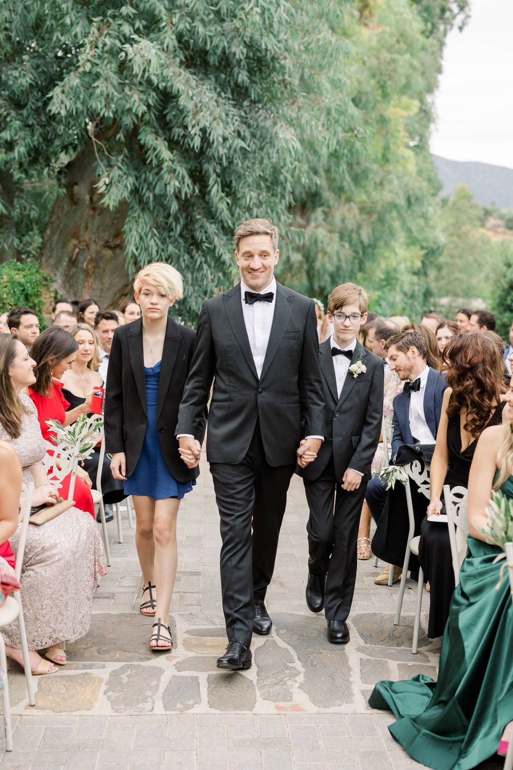 A wedding processional where guests line a stone pathway as attendees walk between them in formal attire.