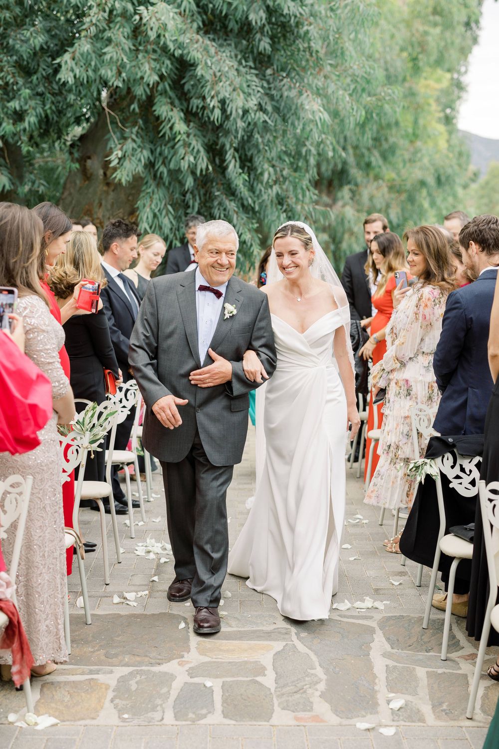 Wedding processional on stone pathway with guests seated on white chairs amid lush greenery.