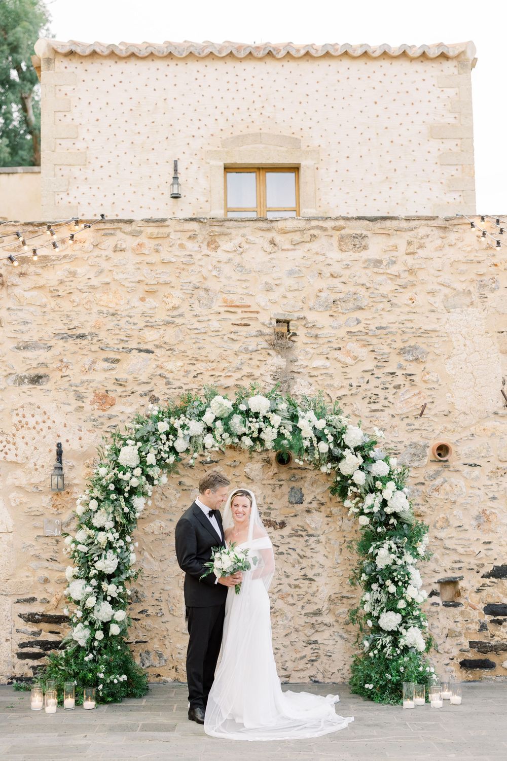 Wedding portrait under floral arch against rustic stone wall.