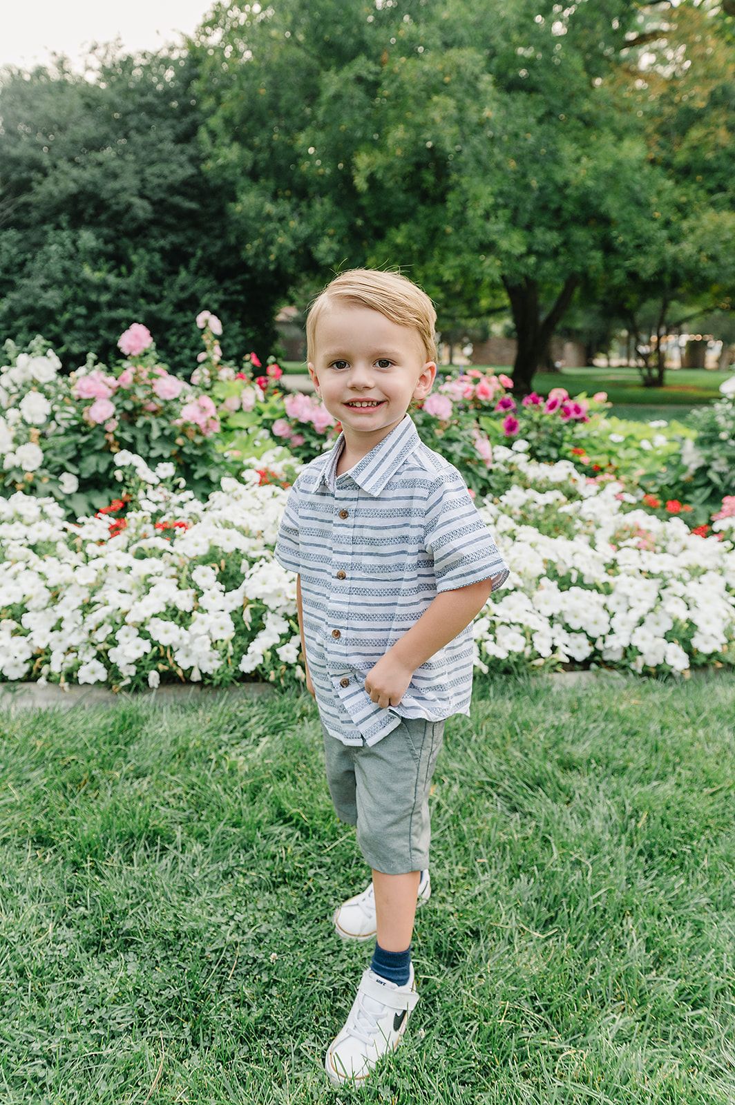 A young child poses happily in front of a garden filled with white and pink flowers.