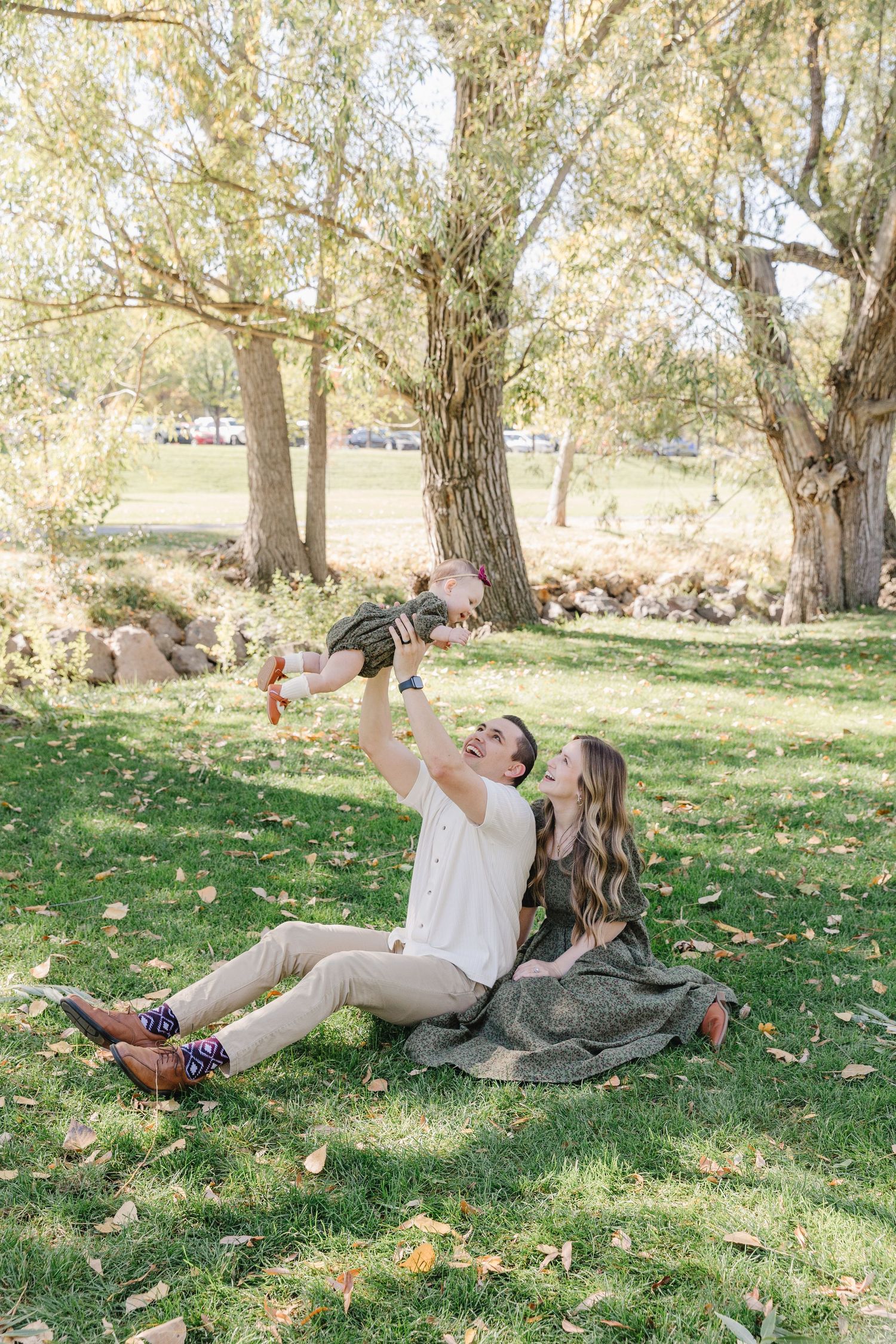 A couple relaxes on grass under willow trees while lifting a baby in a playful moment on a sunny day.