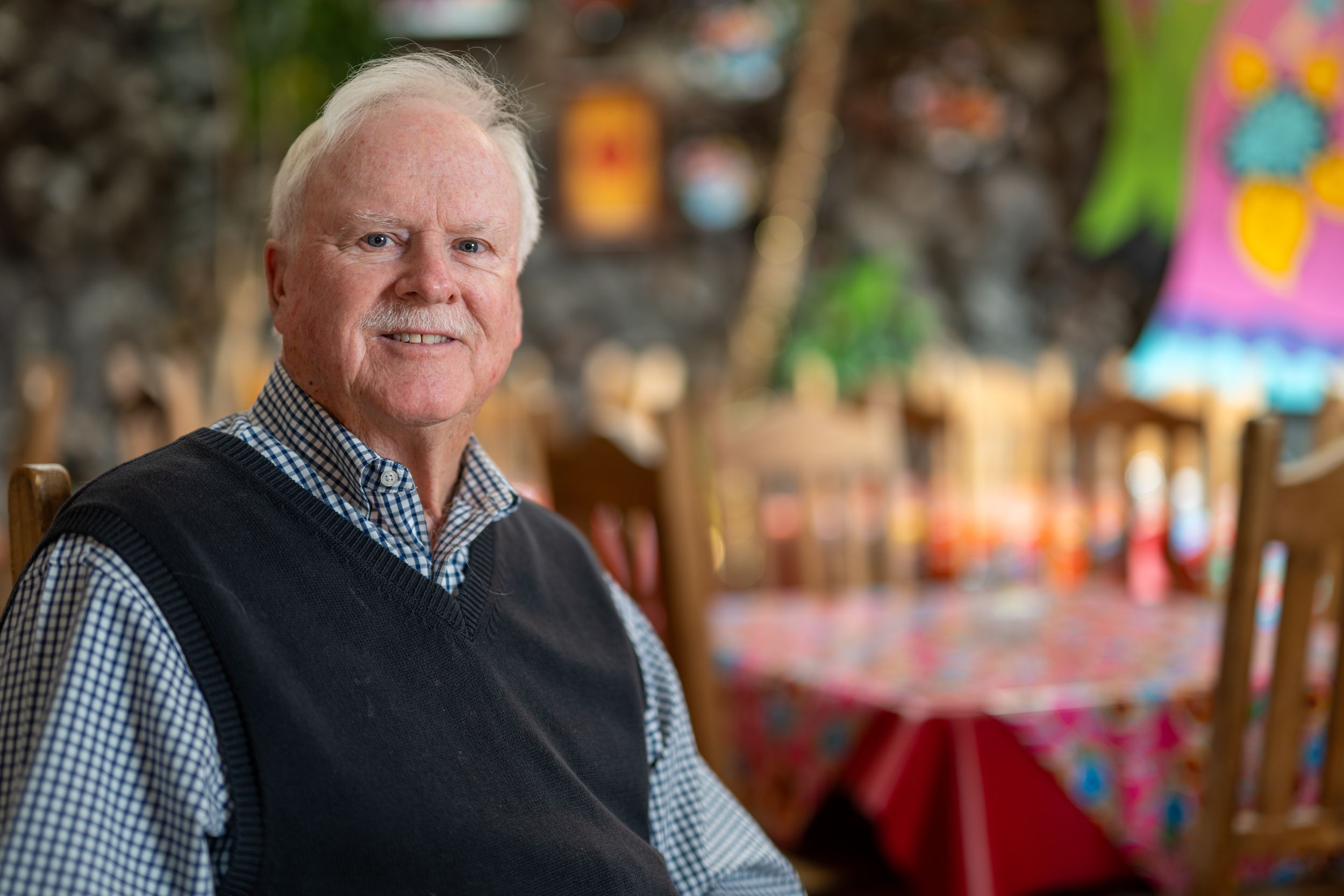 Smiling older man with white hair wearing a navy sweater vest over a checkered shirt, seated in a colorful restaurant with decorated tables.