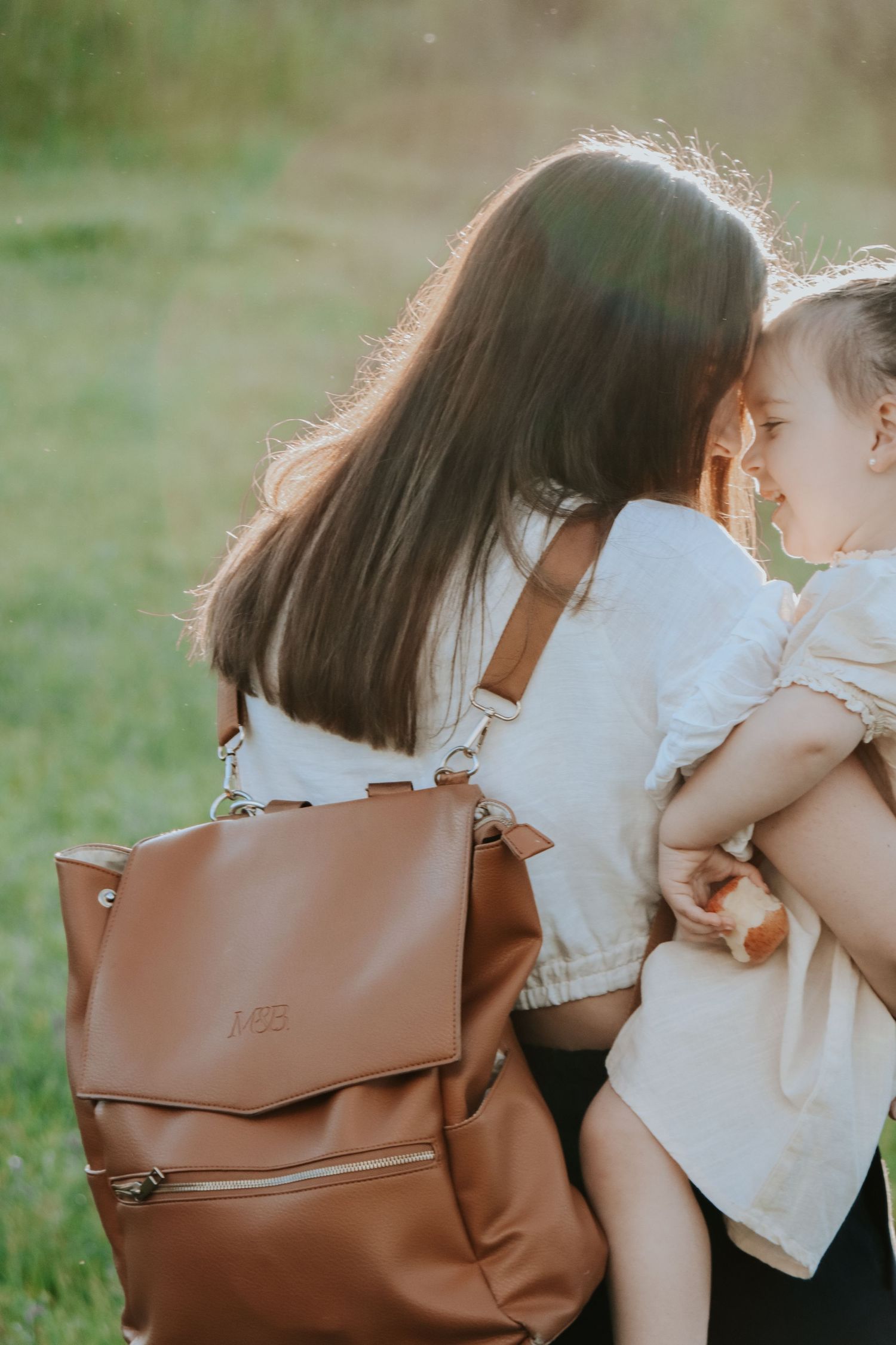 Mother holding toddler while wearing a tan leather monogrammed backpack outdoors in a sunlit green field.