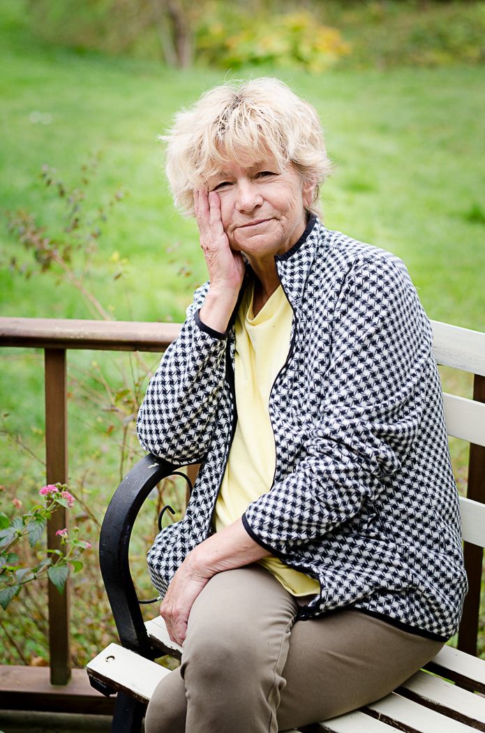 A older woman sitting on her deck smiling with her head in her hand.