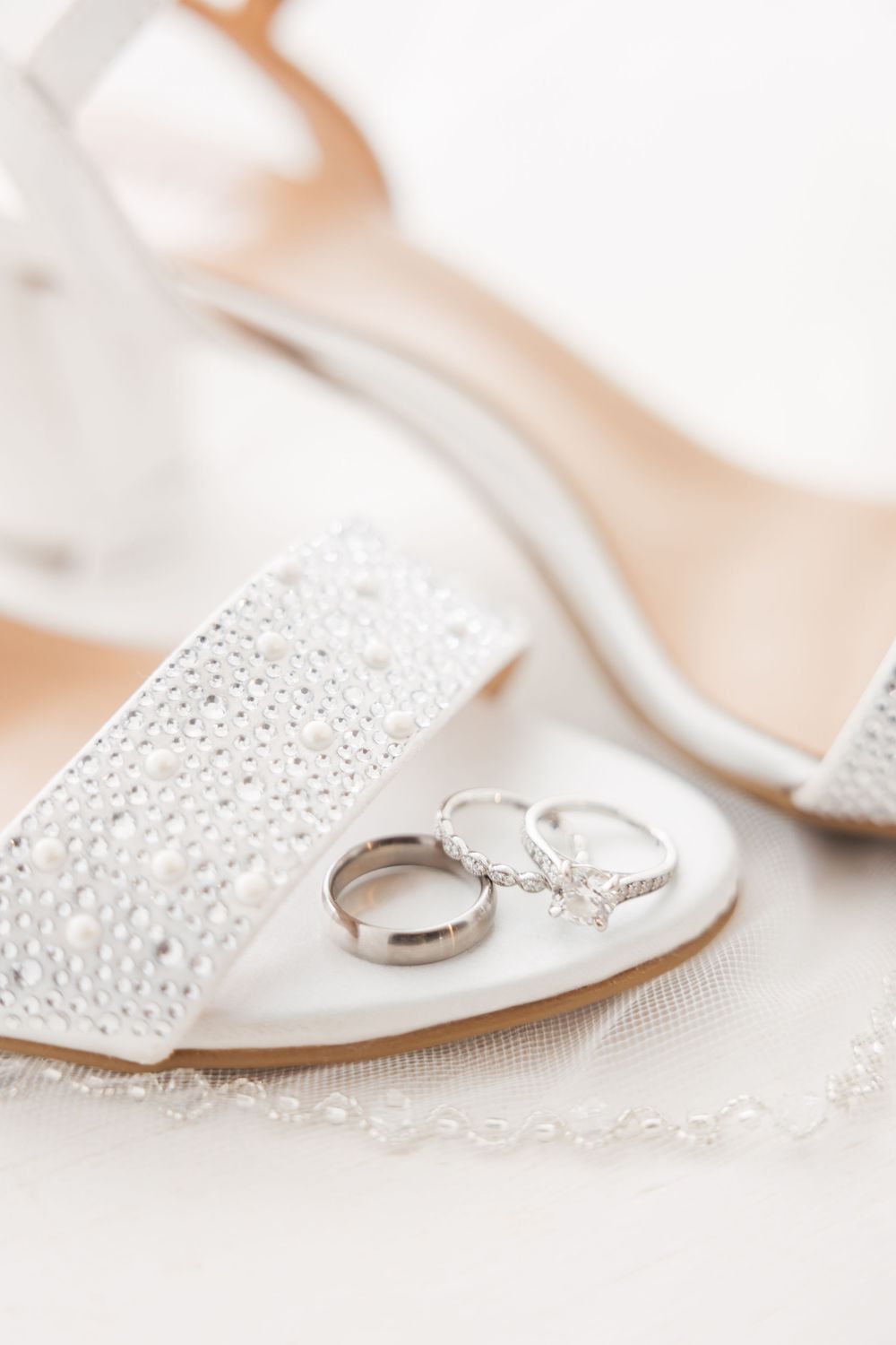 Wedding rings placed on elegant white lace bridal shoe with crystal embellishments and delicate details.