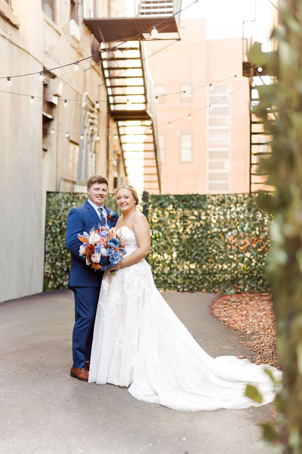 Bride and groom share romantic moments in a covered outdoor walkway decorated with string lights and greenery.