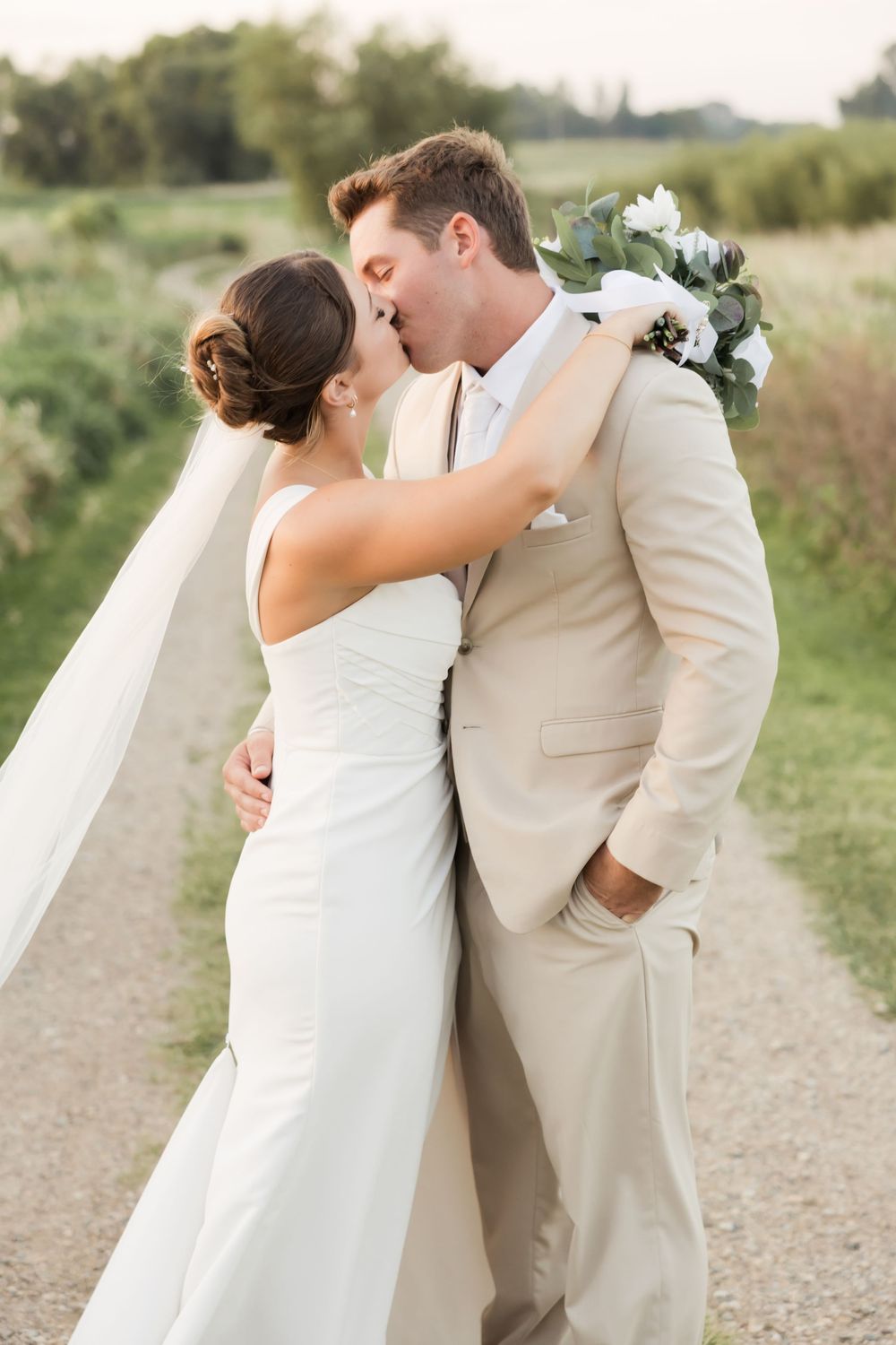 A romantic wedding couple embraces and kisses on a rustic dirt path in a scenic outdoor setting.