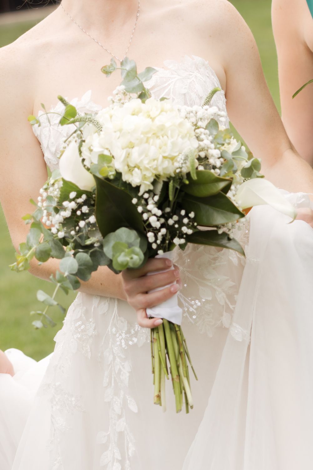 A delicate white wedding bouquet with hydrangeas, baby's breath, and eucalyptus held against a lace wedding dress.