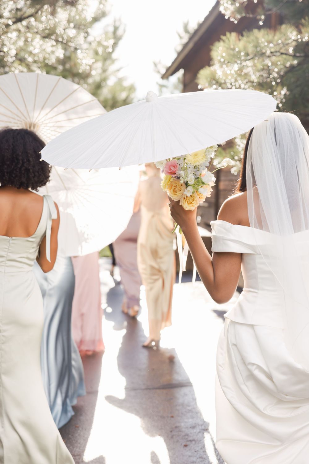 Bridal party with white parasols gathered outdoors in soft sunlight.