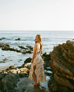 A person in a floral dress stands on rocky coastal cliffs at sunset overlooking the ocean.
