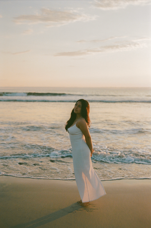 A woman in a white dress stands on a beach during sunset, with gentle waves rolling onto the shore.