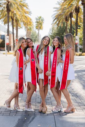 Group wearing matching red graduation stoles pose together on a palm tree-lined walkway in bright sunlight.