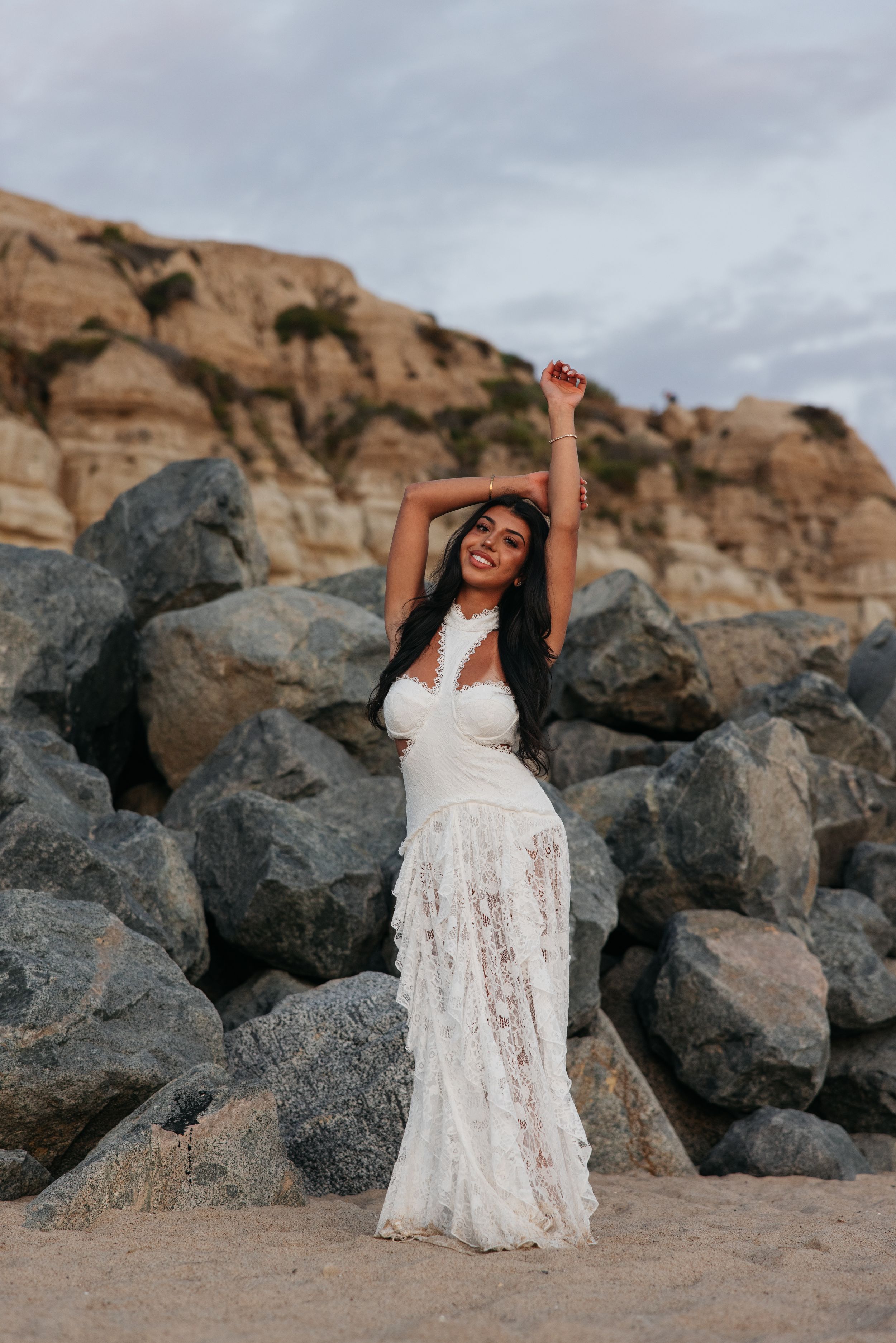 A person in a flowing white lace dress poses gracefully against dark rocks with red cliffs in the background.