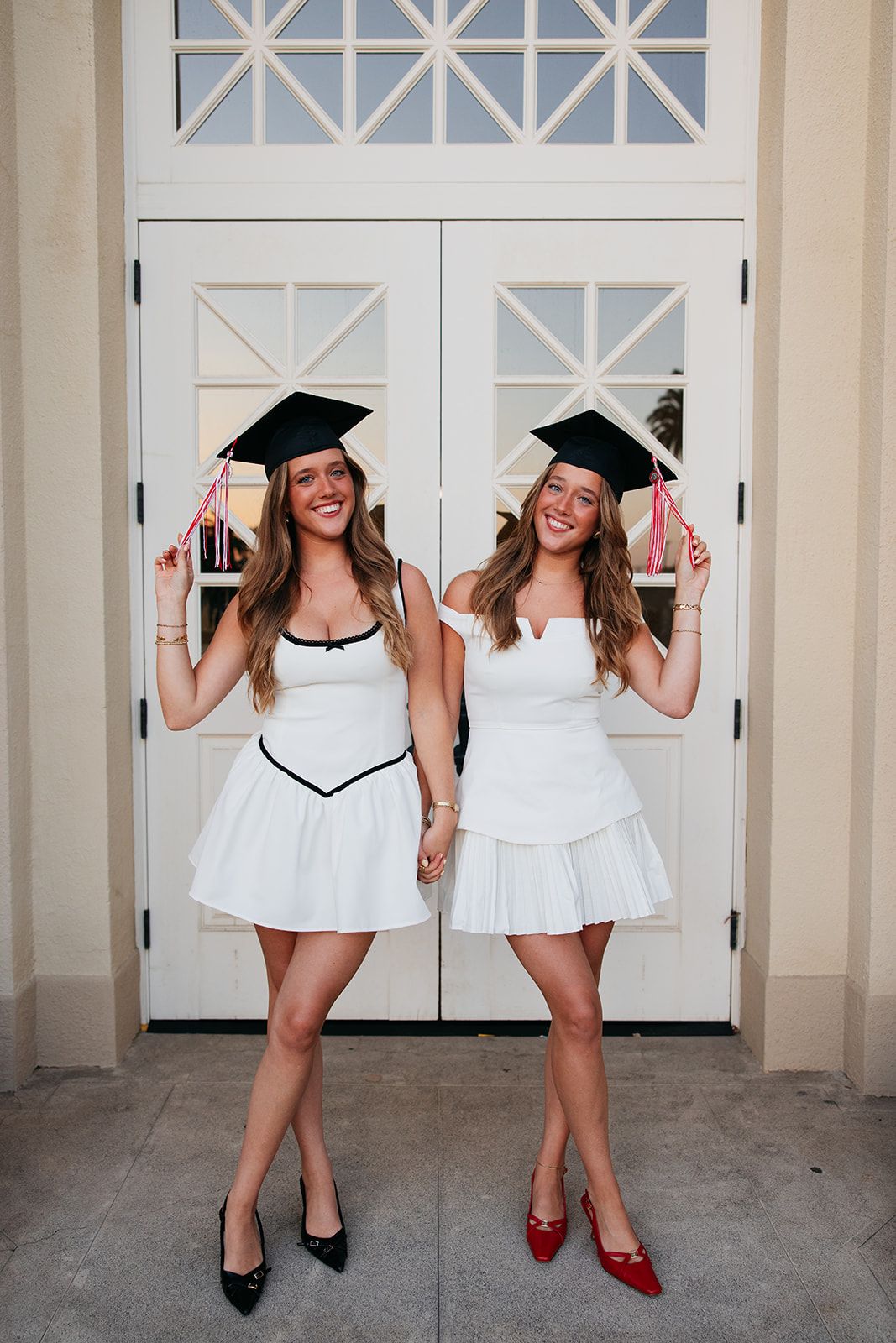 Two graduates in white dresses and graduation caps strike playful poses in front of white double doors.