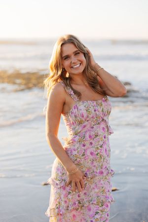 Woman in pink floral sundress poses playfully on beach at sunset.