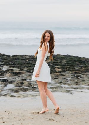 Someone wearing a white sundress poses playfully on a rocky beach shore at sunset with ocean waves in the background.