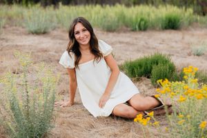 A person in a white dress sits on the ground surrounded by yellow wildflowers and desert sage brush in a natural setting.