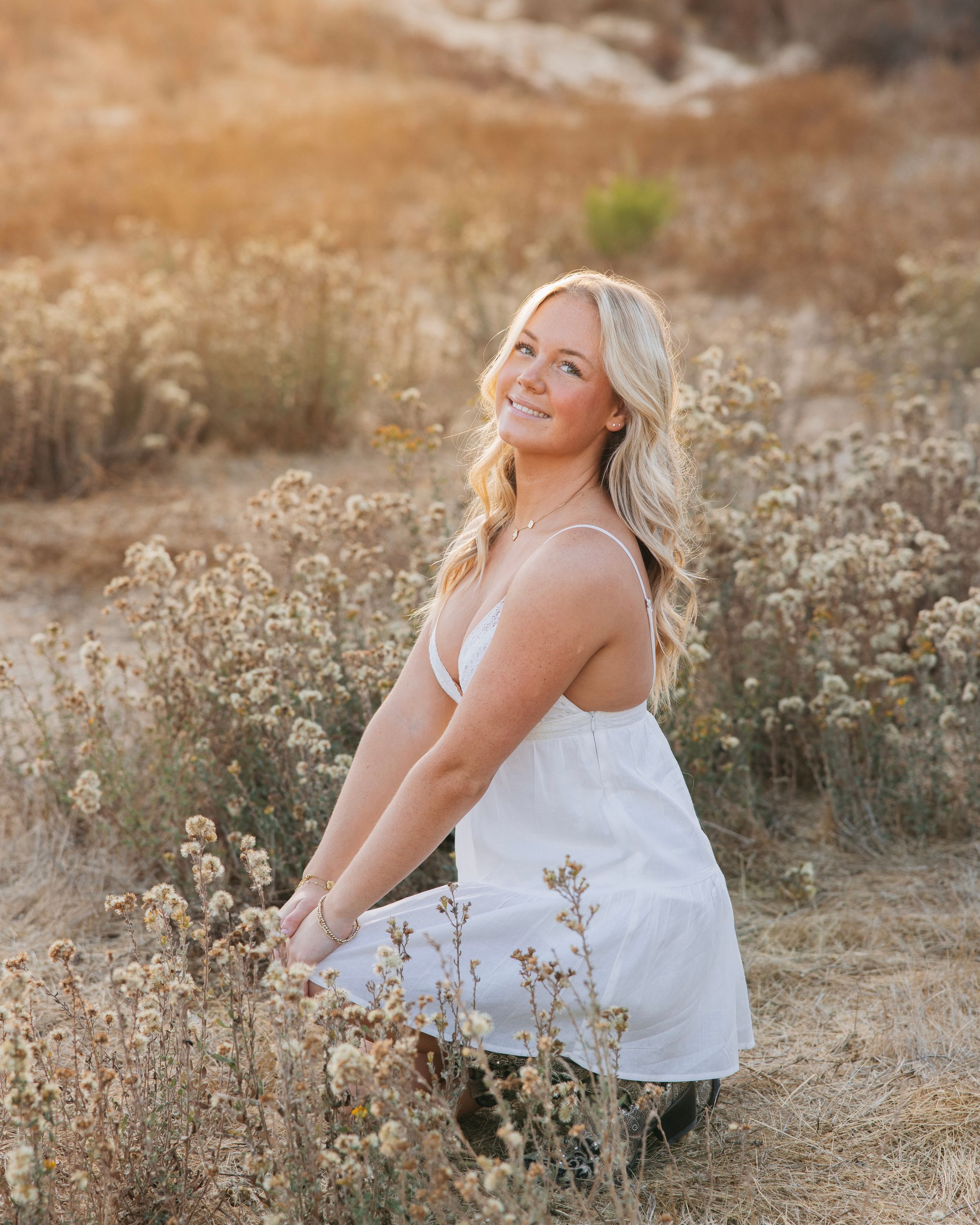 A person in a white top poses joyfully among dried wildflowers during golden hour in a rustic outdoor setting.