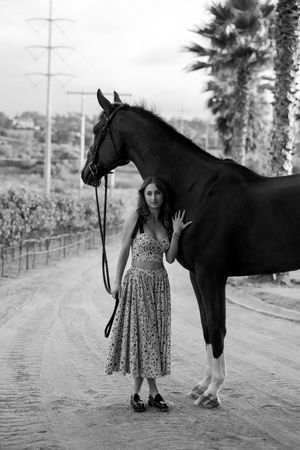 A black horse stands on a dirt road next to someone in a floral dress, with palm trees visible in the background.