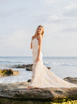 A person in an elegant white lace wedding dress poses on coastal rocks at sunset with ocean waves in the background.