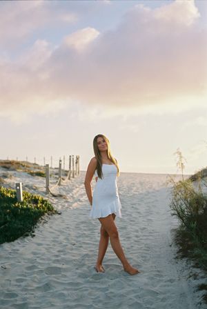 A figure in a flowing white sundress walks barefoot along a sandy beach path at sunset.