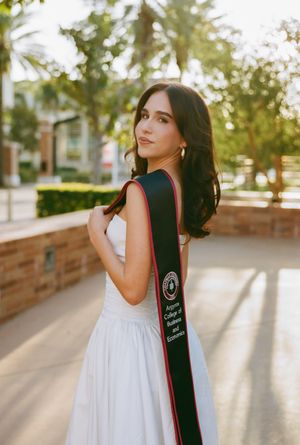 Graduate in white dress poses with graduation stole in warm sunlight on campus pathway.