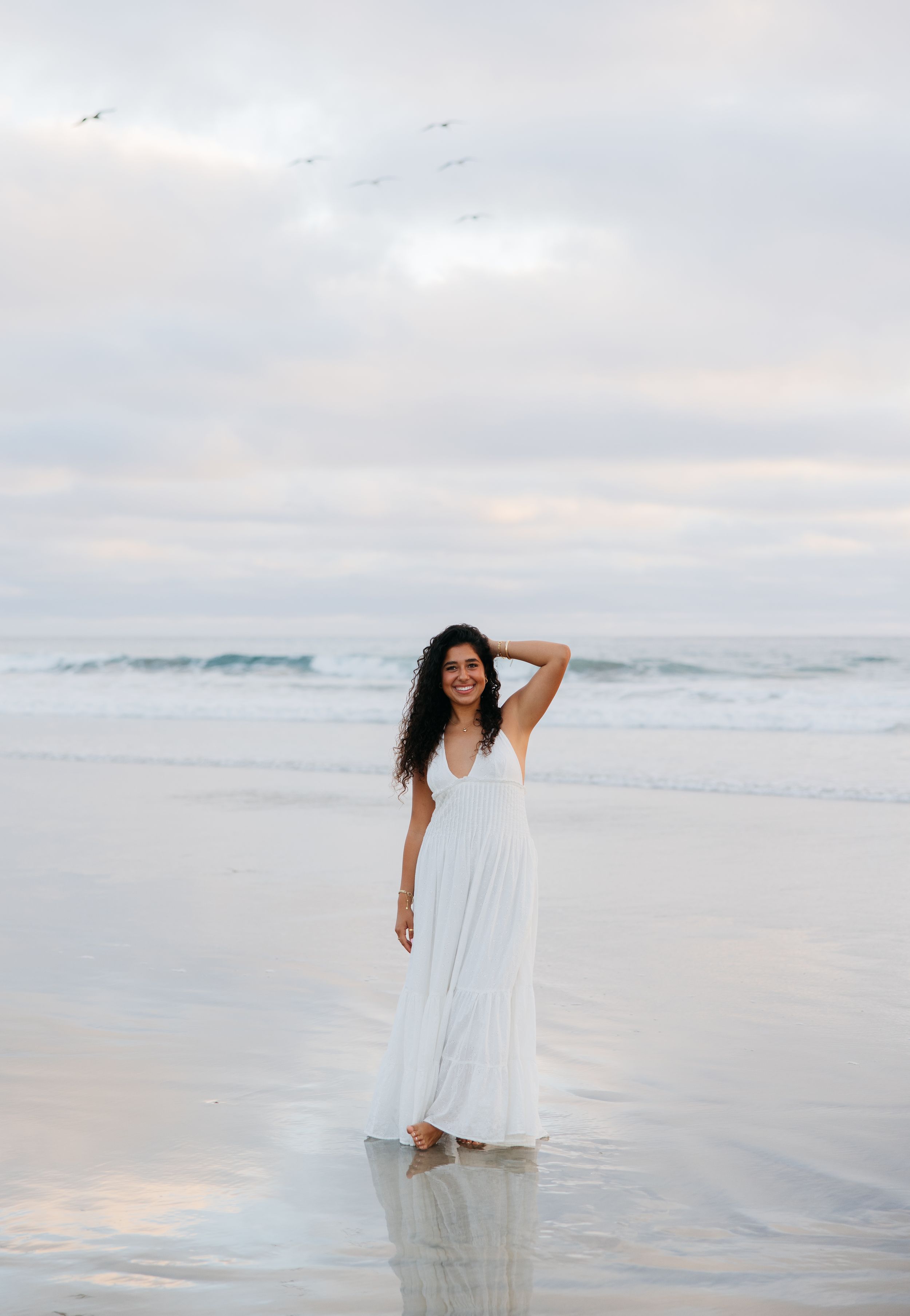 A figure in a flowing white dress stands at the edge of the ocean during sunset, creating a dreamy coastal scene.