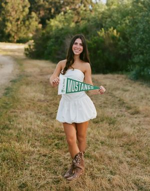 Person in white dress holding a Mustangs sign poses on a dirt path with green foliage in the background.