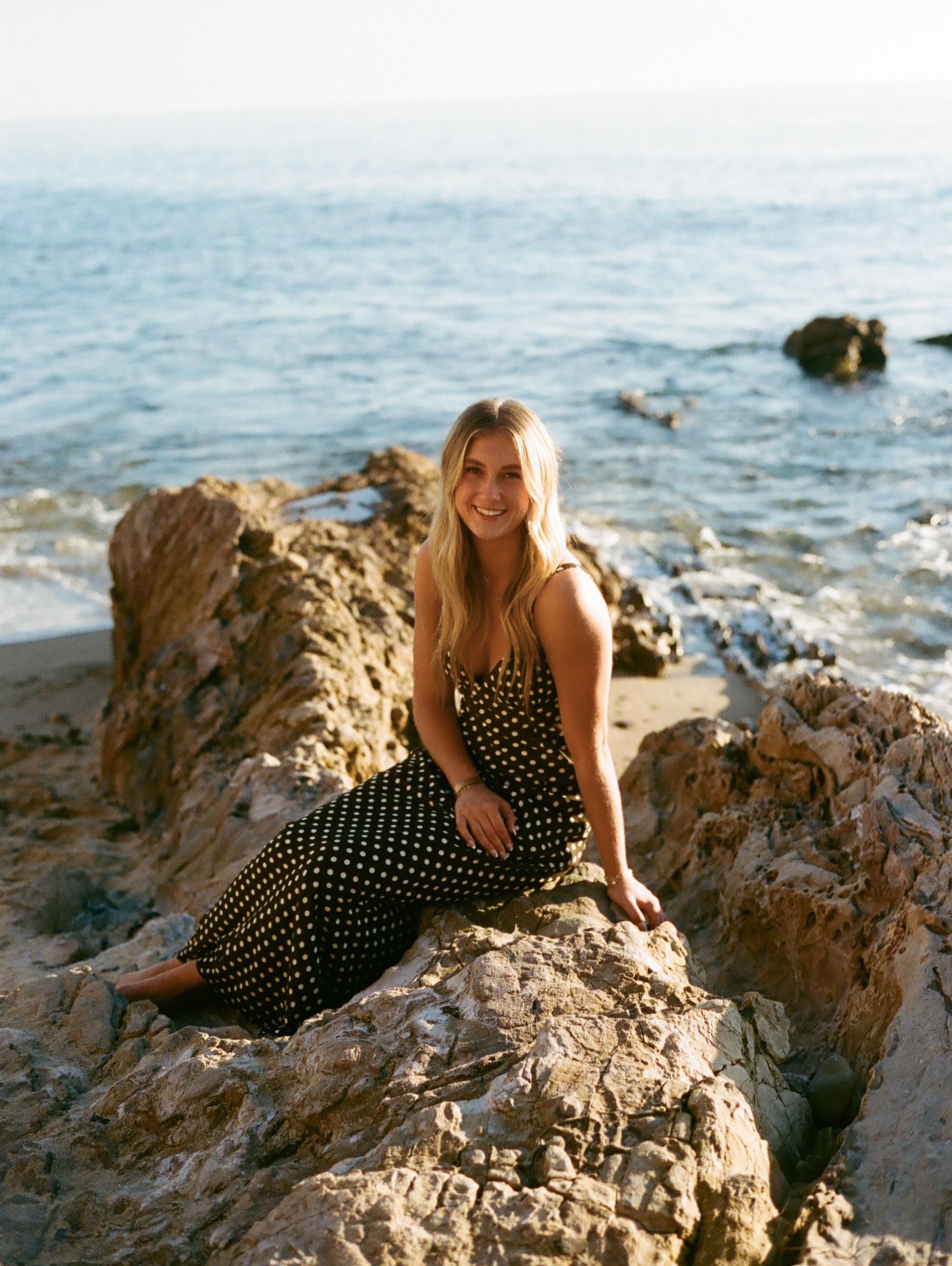 Person in a polka dot dress sits on coastal rocks by the ocean on a sunny day.