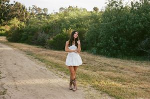 A person in a flowy white dress walks along a golden path during sunset with green trees in the background.