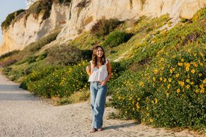 Yellow wildflowers bloom along a coastal cliff path with sandstone bluffs in the background on a sunny day.
