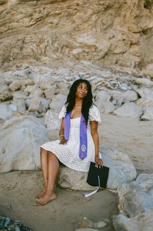 A person wearing a polka dot dress and purple vest relaxes against rocky terrain at the beach.