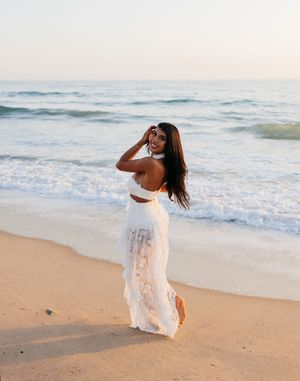 A woman in a white lace dress poses gracefully on a sandy beach with ocean waves in the background.