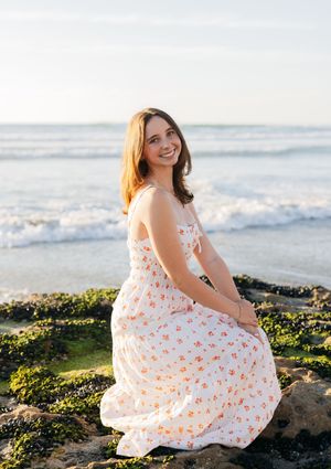 A person in a floral dress sits on rocky beach terrain at sunset with ocean waves in the background.